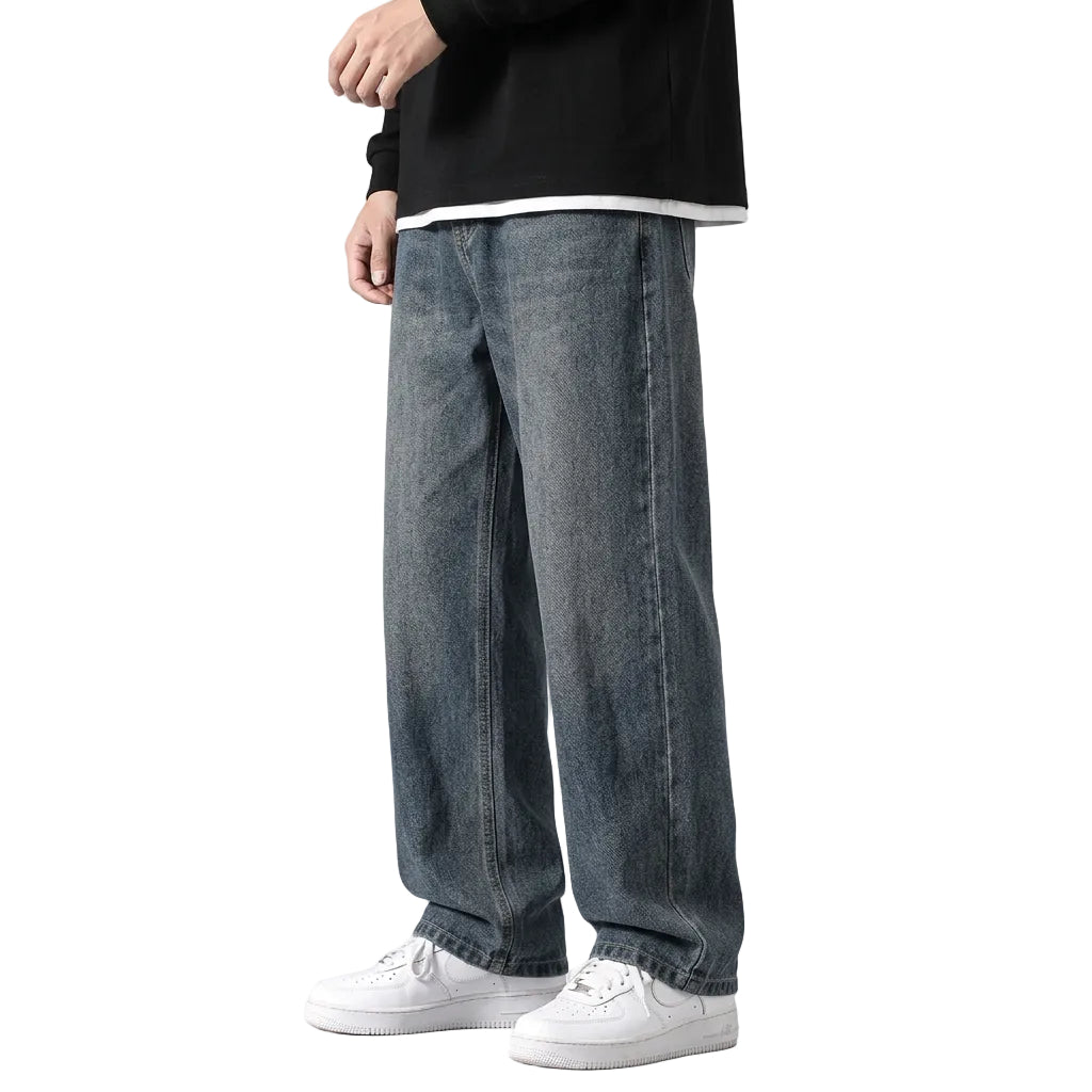 A person wearing loose-fit blue denim jeans with a black oversized top and white sneakers, standing against a plain gray wall.