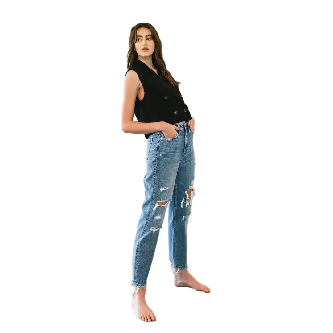 Woman wearing a black sleeveless top and ripped blue jeans standing against a white wall.