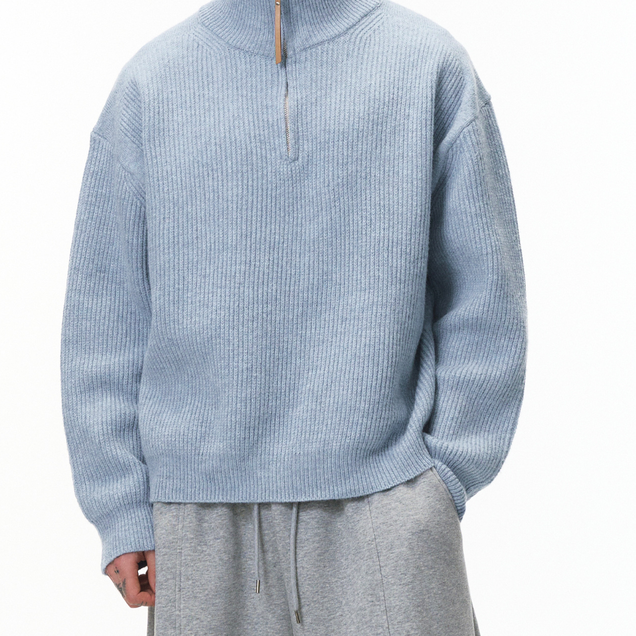 A person wearing a light blue ribbed knit half-zip sweater with a small gold-toned zipper pull, paired with gray drawstring sweatpants, photographed against a plain white studio background.