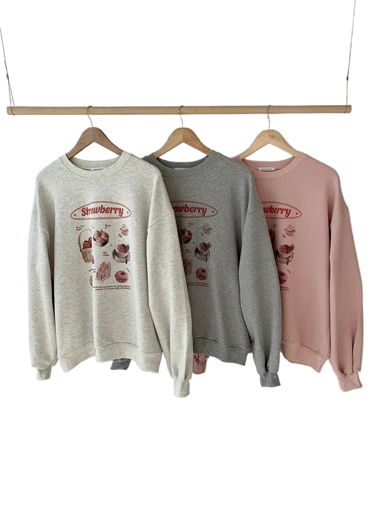 Three crewneck sweatshirts in heather cream, heather gray, and dusty pink, hanging on wooden hangers from a wooden clothes rack against a plain light gray wall. Each sweatshirt features a strawberry-themed graphic print on the front.