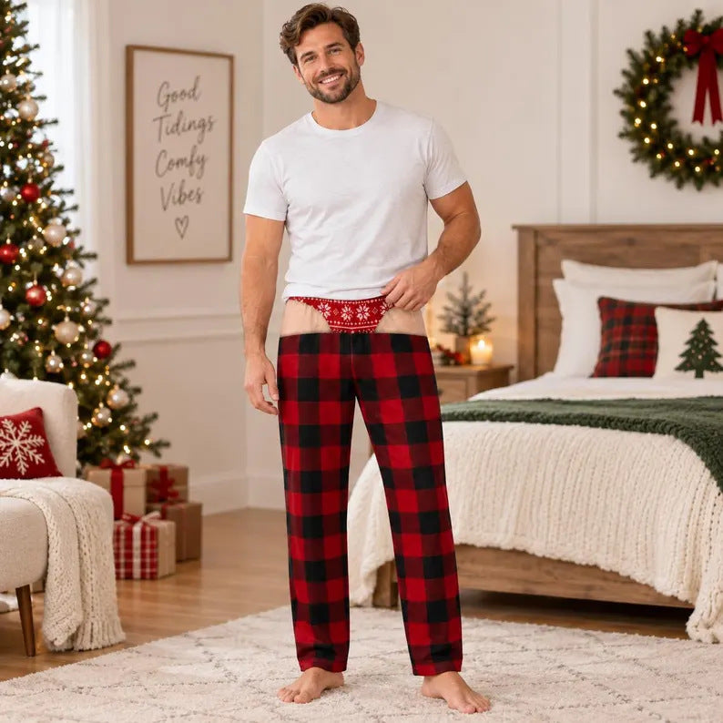 A man stands in a decorated bedroom wearing red and black plaid Christmas pajama pants with a red patterned waistband, paired with a white t-shirt. The room features a Christmas tree, a bed with holiday pillows, and festive wall decorations.