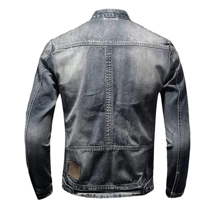 A dark gray washed denim jacket shown from the back, featuring a central seam, decorative stitching, and a small brown leather patch near the lower left back, set against a dark gray gradient background.