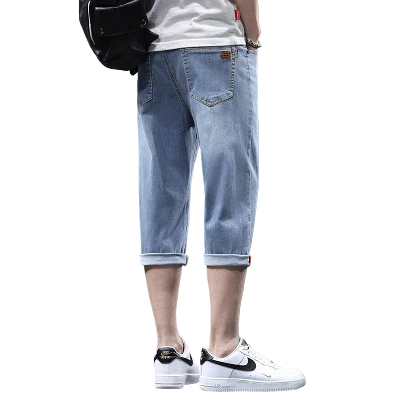 A person shown from the back wearing light blue 3/4-length denim jeans with rolled cuffs, a white T-shirt, a black backpack, and white sneakers against a dark gray wall.