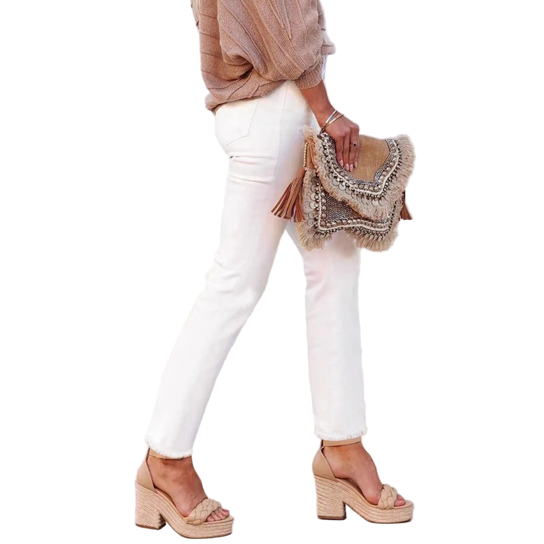 A person wearing white pants and beige platform sandals leans against a white wall, holding a woven fringe clutch bag with tassels.
