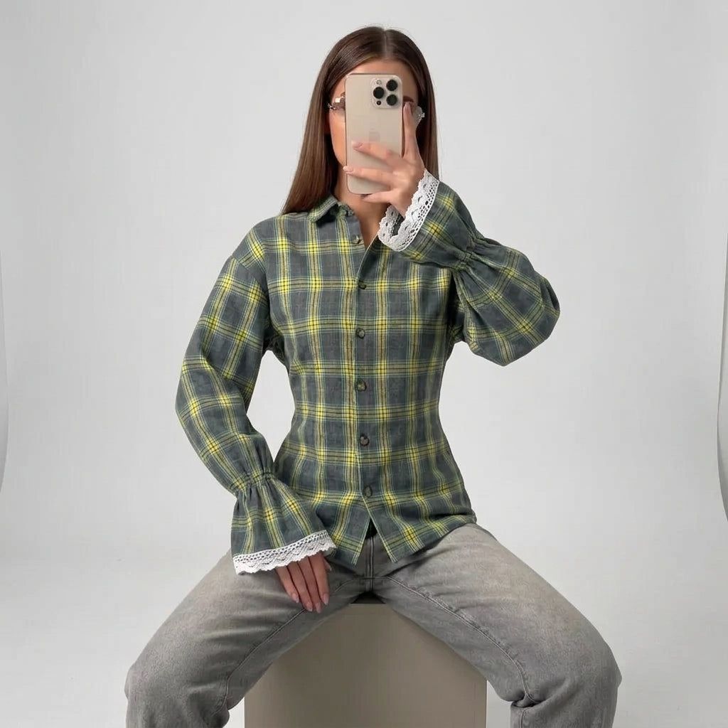 A woman takes a mirror selfie wearing a green and yellow plaid button-up shirt with white lace trim on the cuffs, paired with gray jeans, sitting on a light gray studio pedestal.