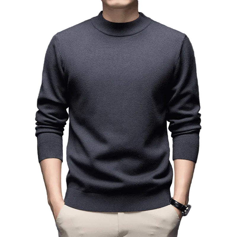 A man wearing a dark charcoal gray mock neck knit sweater and light beige trousers, shown from the chin down against a dark gray studio background.