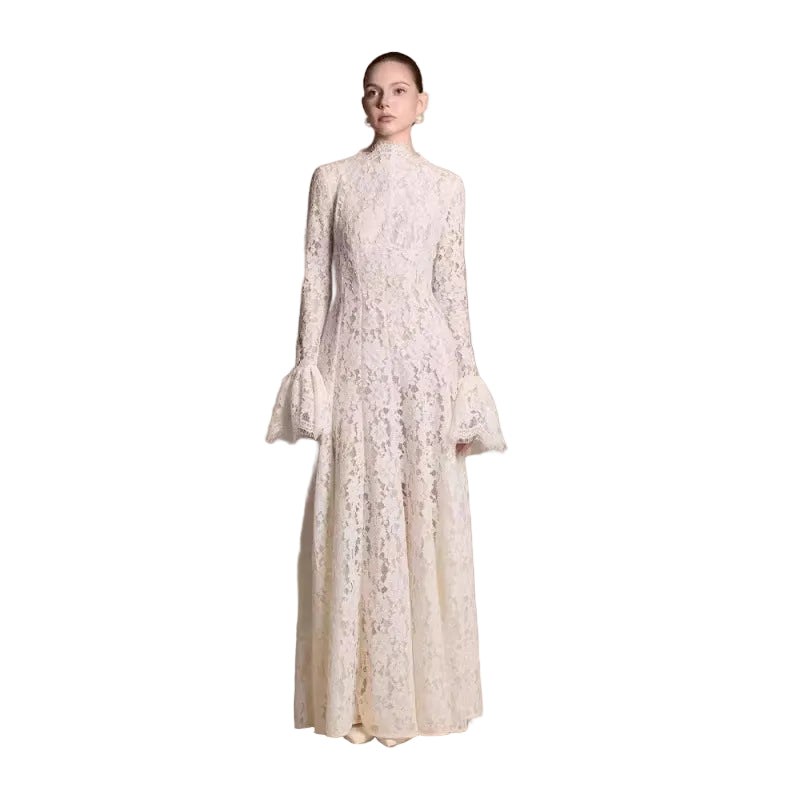 A model stands against a plain beige studio backdrop wearing a long-sleeve, high-neck lace maxi dress in a warm ivory or cream tone.