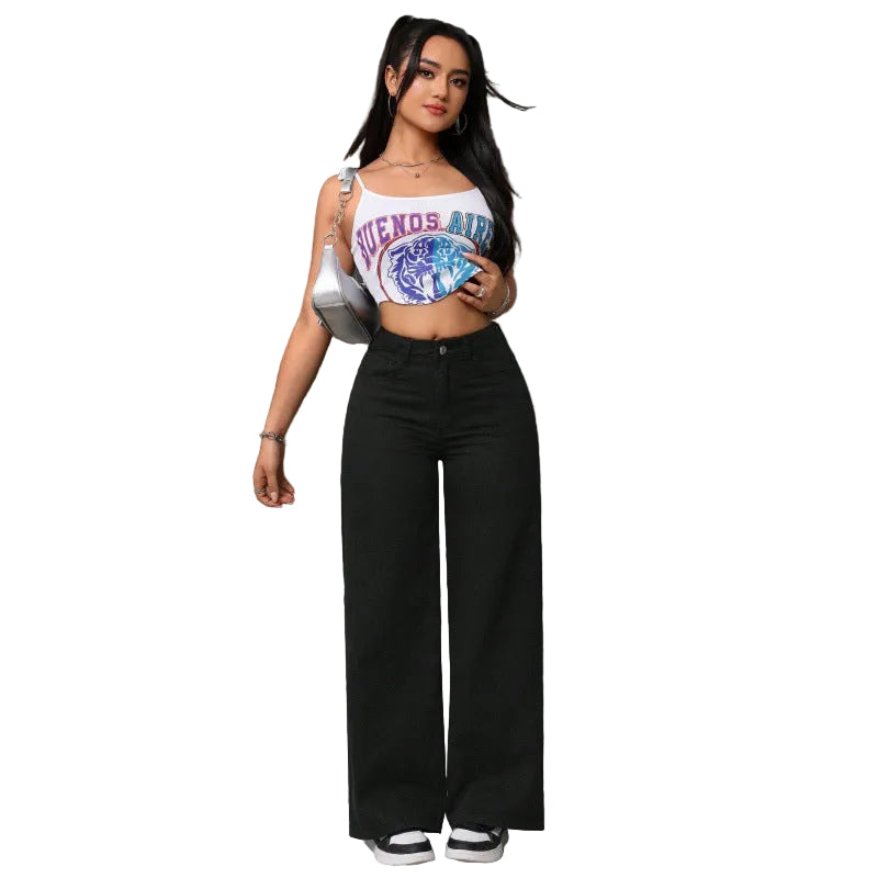 A woman stands in a concrete parking garage wearing a white cropped tank top with a colorful graphic print and black wide-leg pants, holding a silver shoulder bag.