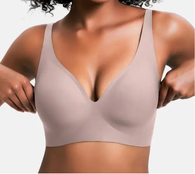 A close-up of a woman wearing a seamless, scoop-neck bra in a light taupe color, with her hands adjusting the straps against a plain white background.