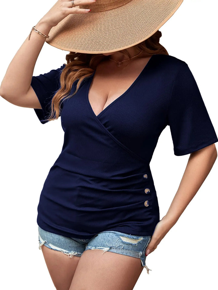 A woman wearing a navy blue short-sleeve wrap-style top with side buttons, paired with denim shorts and a large straw hat, against a plain white background.