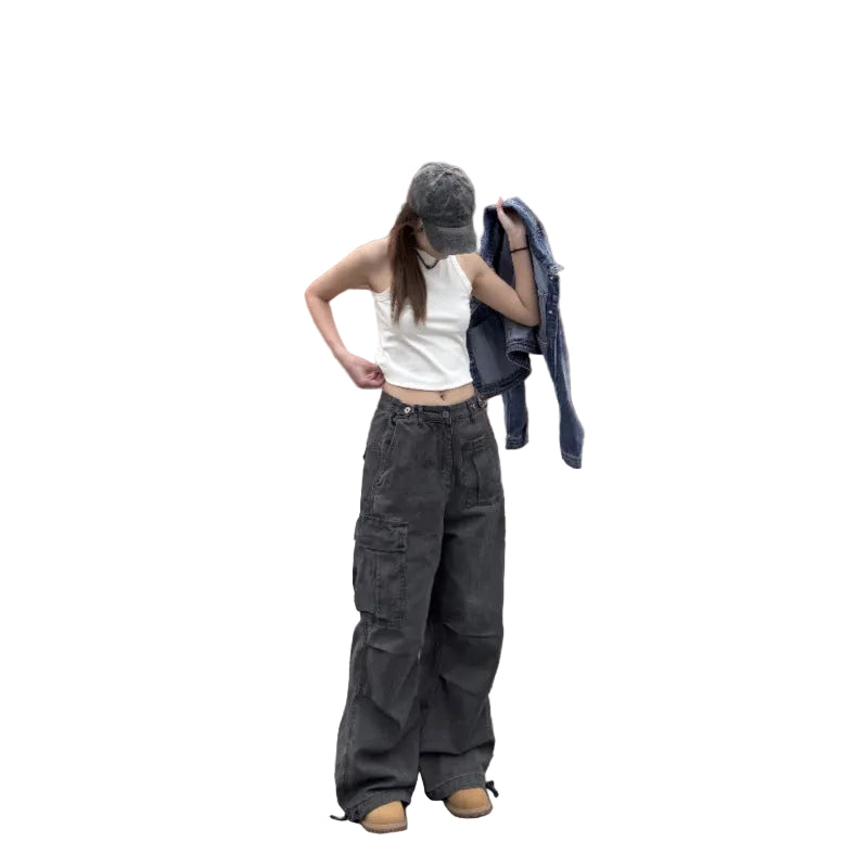 A woman stands on a wooden deck wearing a white ribbed tank top, dark gray wide-leg cargo pants, a gray baseball cap, and tan shoes, while holding a blue denim jacket against a red-brown slatted wall.