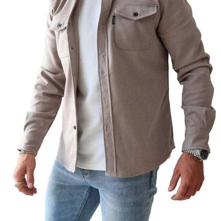 Person wearing a beige jacket over a white shirt and blue jeans on a plain background