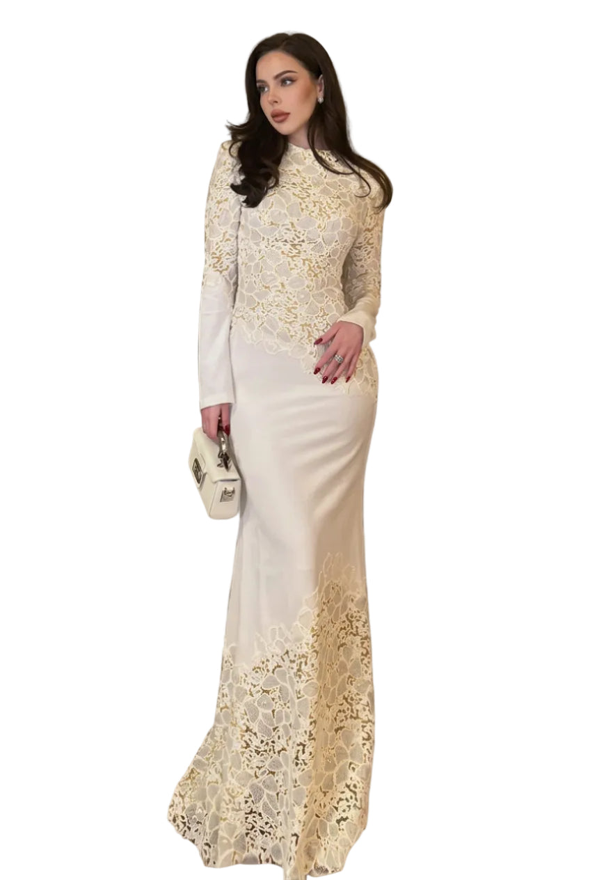 A woman wearing a long-sleeve, high-neck, off-white lace and satin mermaid gown stands on a red carpeted staircase, holding a small white handbag.