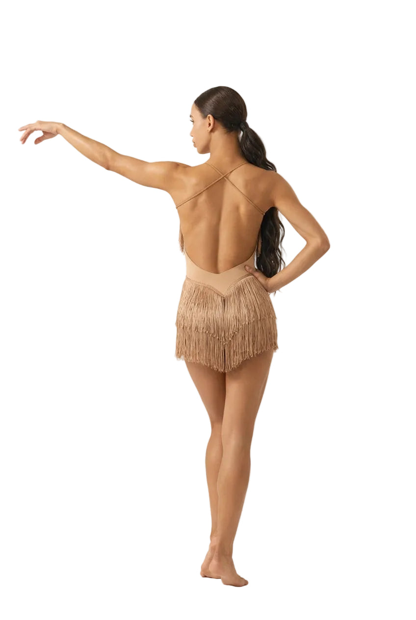 A woman posing from behind wearing a light beige fringed Latin dance leotard with a strappy open back, standing in front of a ballet barre in a bright studio.