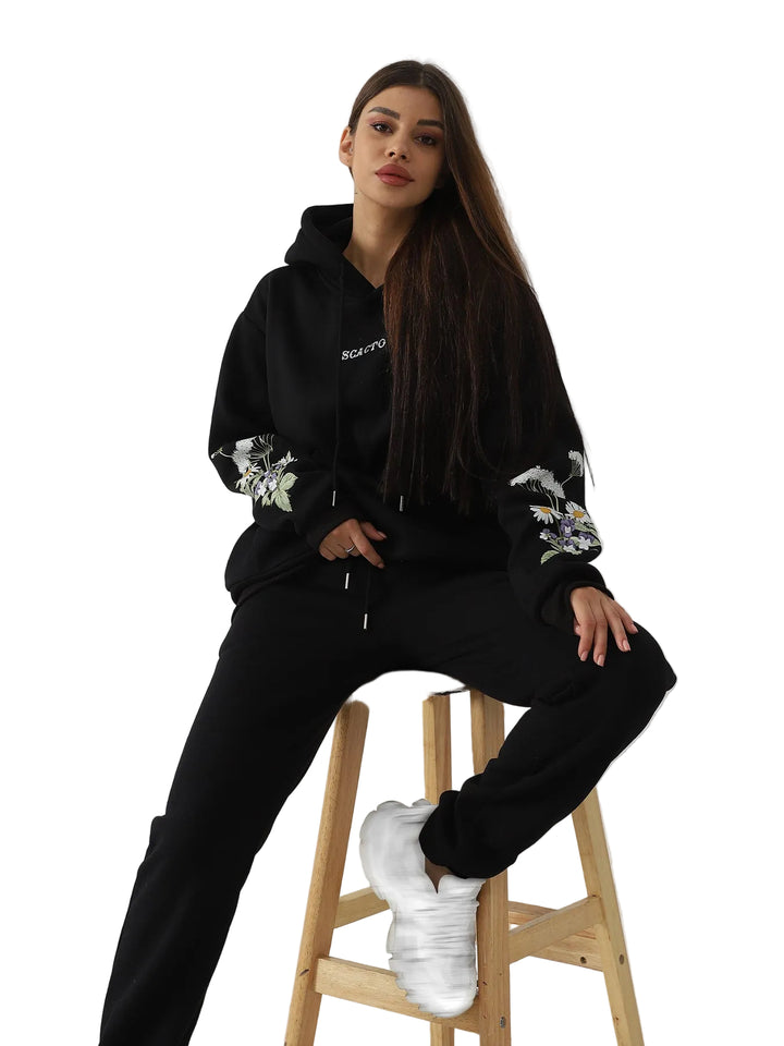 Woman wearing a black hoodie and pants with floral designs, sitting on a wooden stool against a white wall.