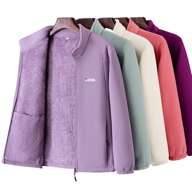Four fleece zip-up jackets hanging on wooden hangers against a light cream background, shown in lavender, sage green, cream, coral pink, and purple. The front lavender jacket is most prominent, with a small white and red logo on the left chest.