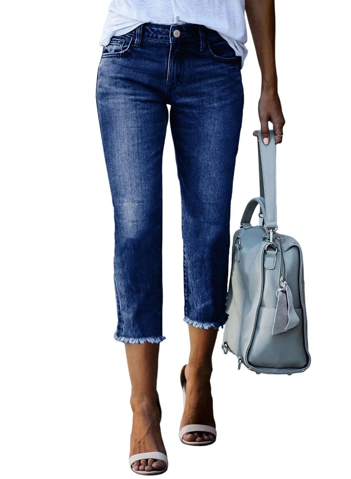 A model wearing dark blue cropped denim jeans with a frayed hem, holding a light blue-gray shoulder bag, against a plain white background.