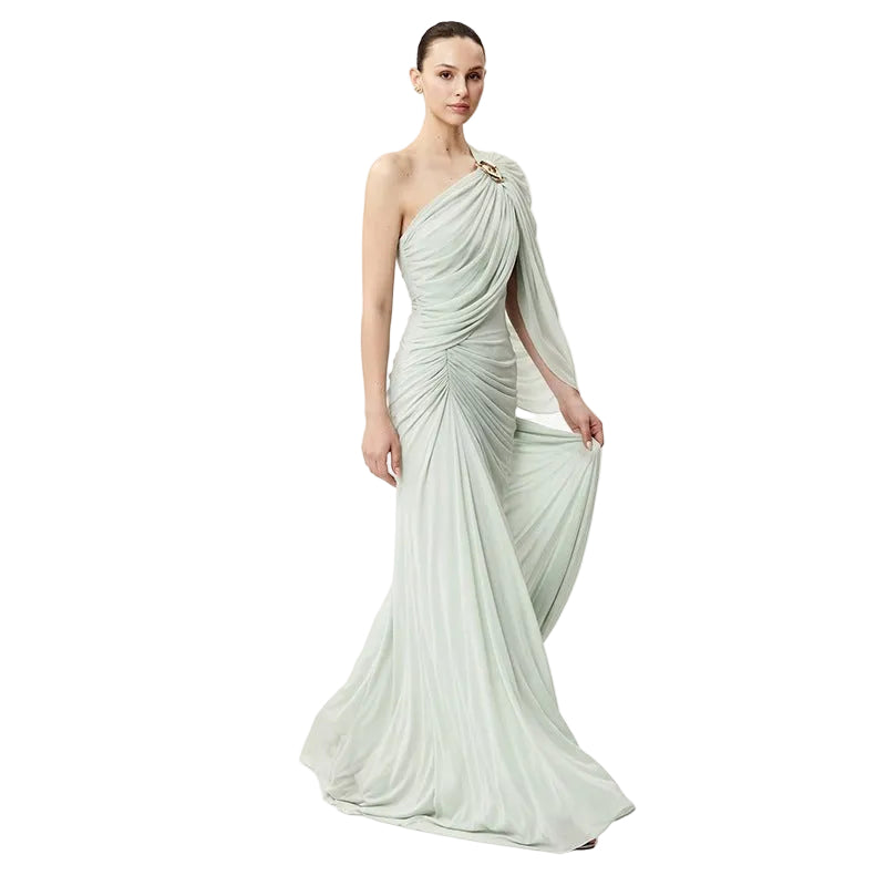 A woman modeling a floor-length draped gown in a pale sage green color, featuring a one-shoulder design with a draped sleeve and gathered bodice, set against a plain white studio background.
