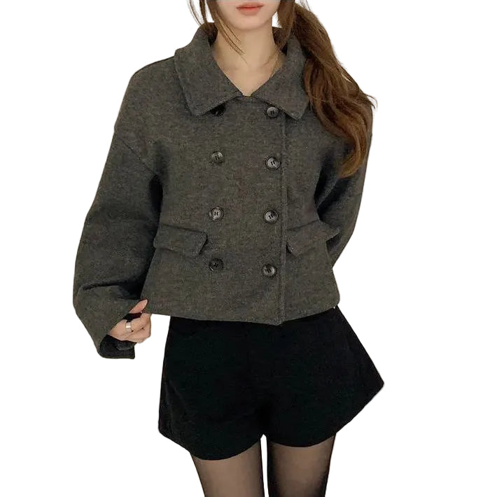 A woman wearing a dark gray double-breasted cropped jacket with dark buttons, paired with black shorts and sheer black tights, standing against a plain beige wall.