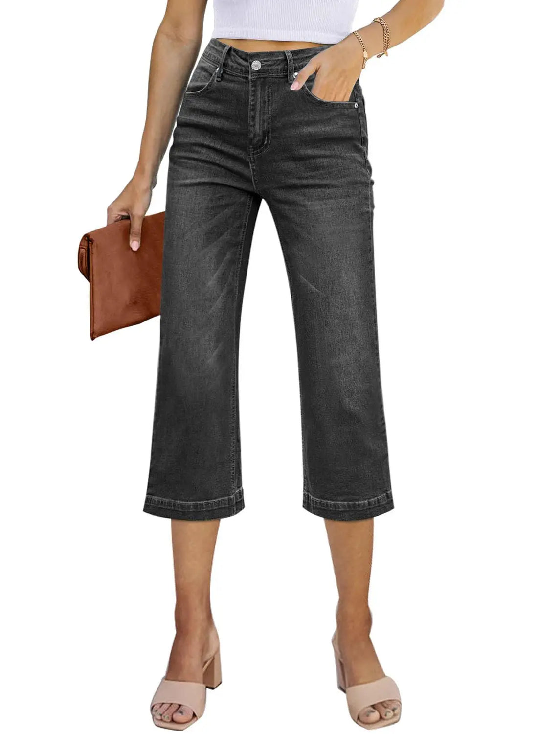 A model wearing dark gray cropped denim jeans, holding a brown clutch, and wearing beige heeled sandals against a plain white background.