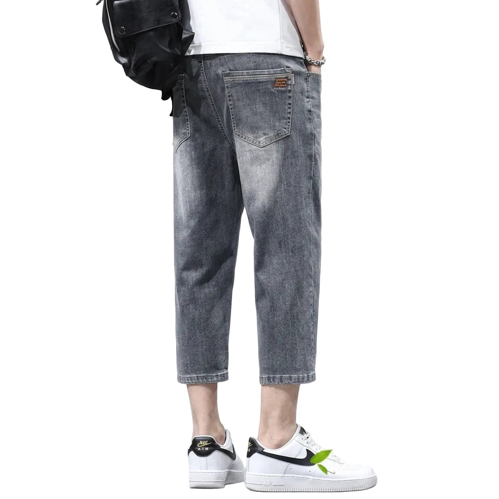 A person wearing faded gray cropped denim jeans with a white t-shirt and white sneakers, shown from the waist down against a dark gray studio background. The image includes promotional text reading 'Thin ice oxygen' and a circular inset close-up of the back pocket area, with decorative ice cubes at the bottom.