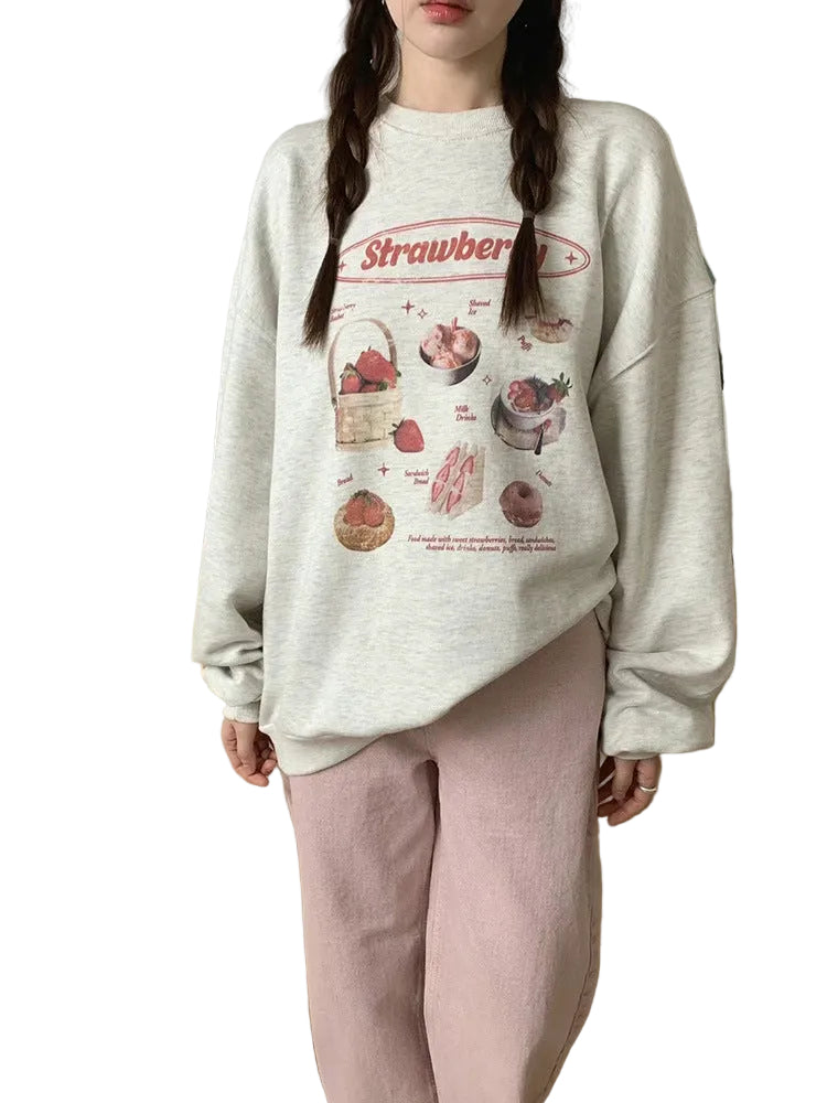 A person wearing an oversized light heather gray sweatshirt with a strawberry-themed graphic print on the front, paired with light pink pants, standing in a kitchen setting.