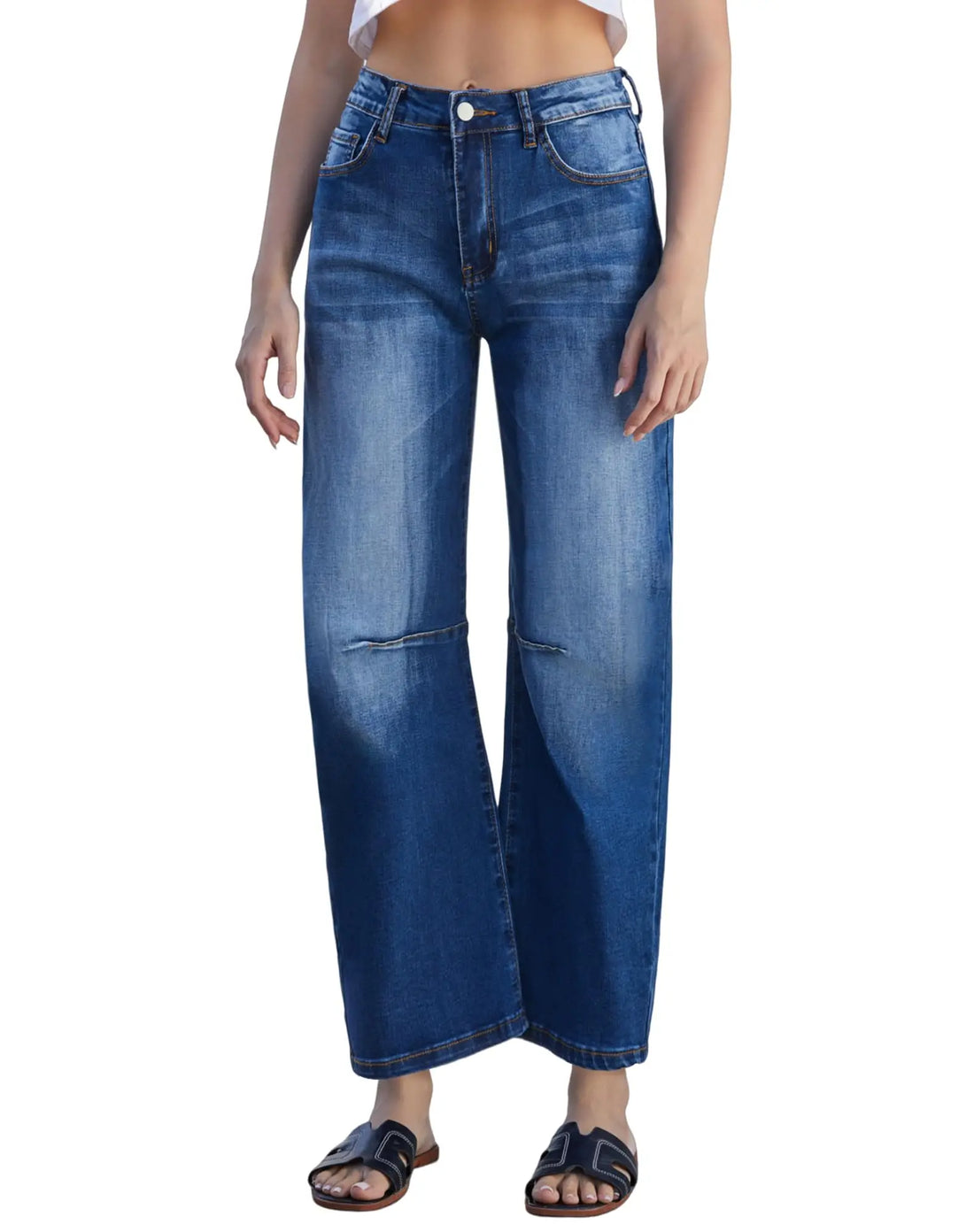 A model wearing a pair of high-waisted, wide-leg cropped blue denim jeans with distressed details and a button closure, paired with black slide sandals against a plain white background.