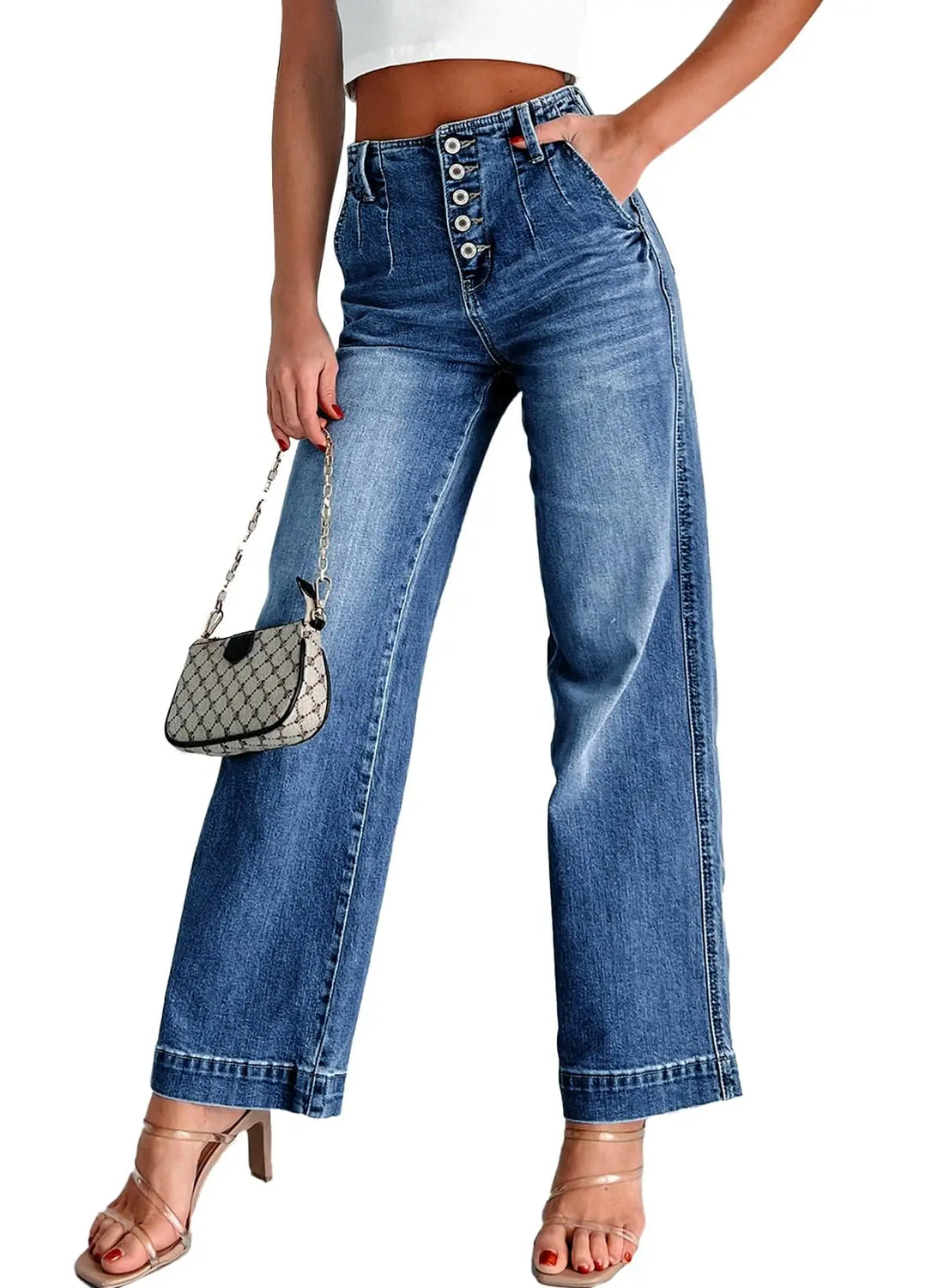 A model wearing high-waisted, wide-leg blue denim jeans with decorative silver buttons down the front fly. The model is also holding a small patterned handbag and wearing clear-strap high-heeled sandals, set against a plain white background.