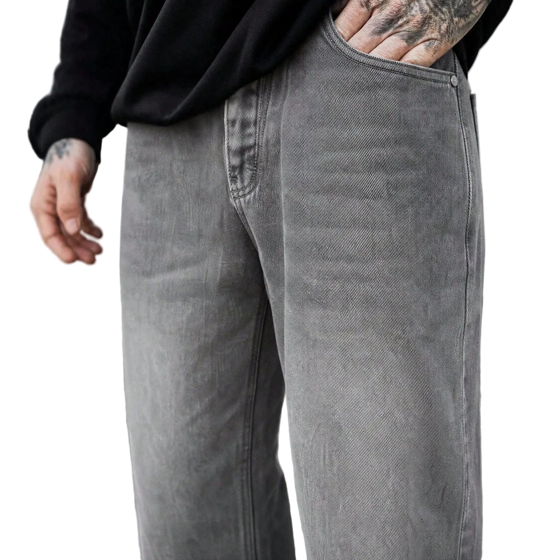 A close-up of a person wearing faded gray denim jeans and a black sweatshirt, with one hand in the pocket and the other hanging by the side. The background shows a blurred outdoor walkway.