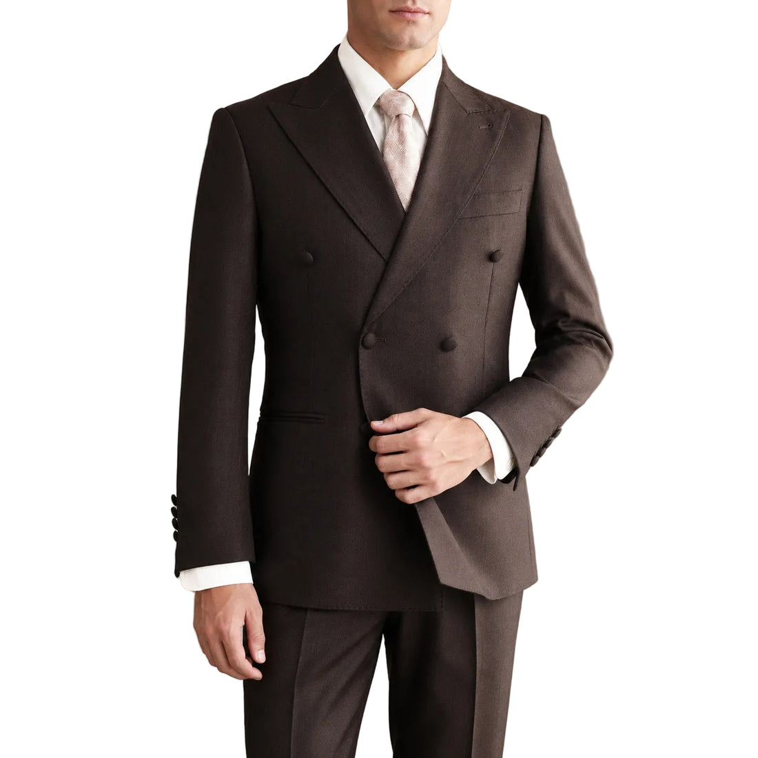 A man wearing a dark brown double-breasted suit with a white dress shirt and a light pink tie, standing in an elegant room with beige walls and dark brown curtains.