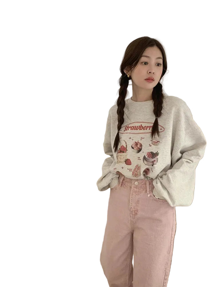 A woman stands indoors wearing a light gray oversized sweatshirt with a strawberry graphic, pink jeans, and black-and-white sneakers. A large mirror reflects her back, showing the full outfit.
