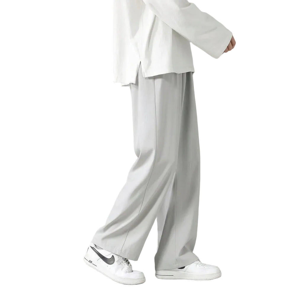 A person shown from the waist down wearing light gray wide-leg pants, a white top, and white sneakers against a muted sage green studio background.