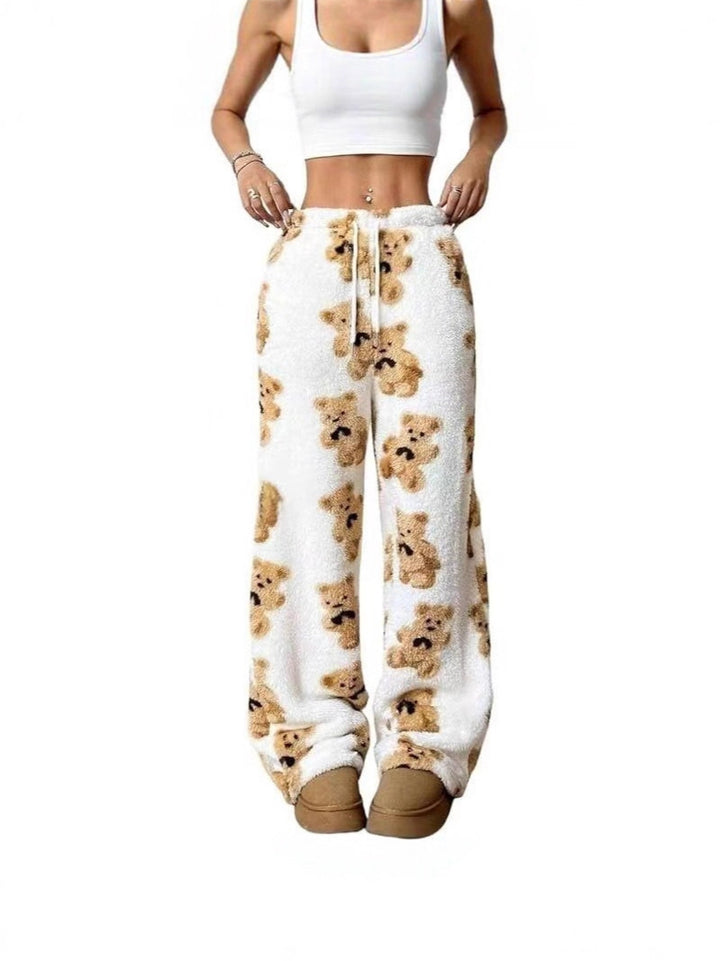 A model wearing white plush fleece sweatpants with an all-over teddy bear print, paired with a white crop top and tan slippers, shown against a plain white background.