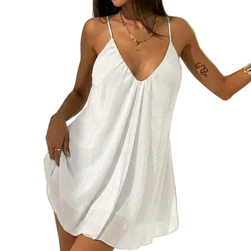 A woman wearing a white sleeveless mini dress with thin straps and a deep V-neckline, standing against a light wall with soft shadows.