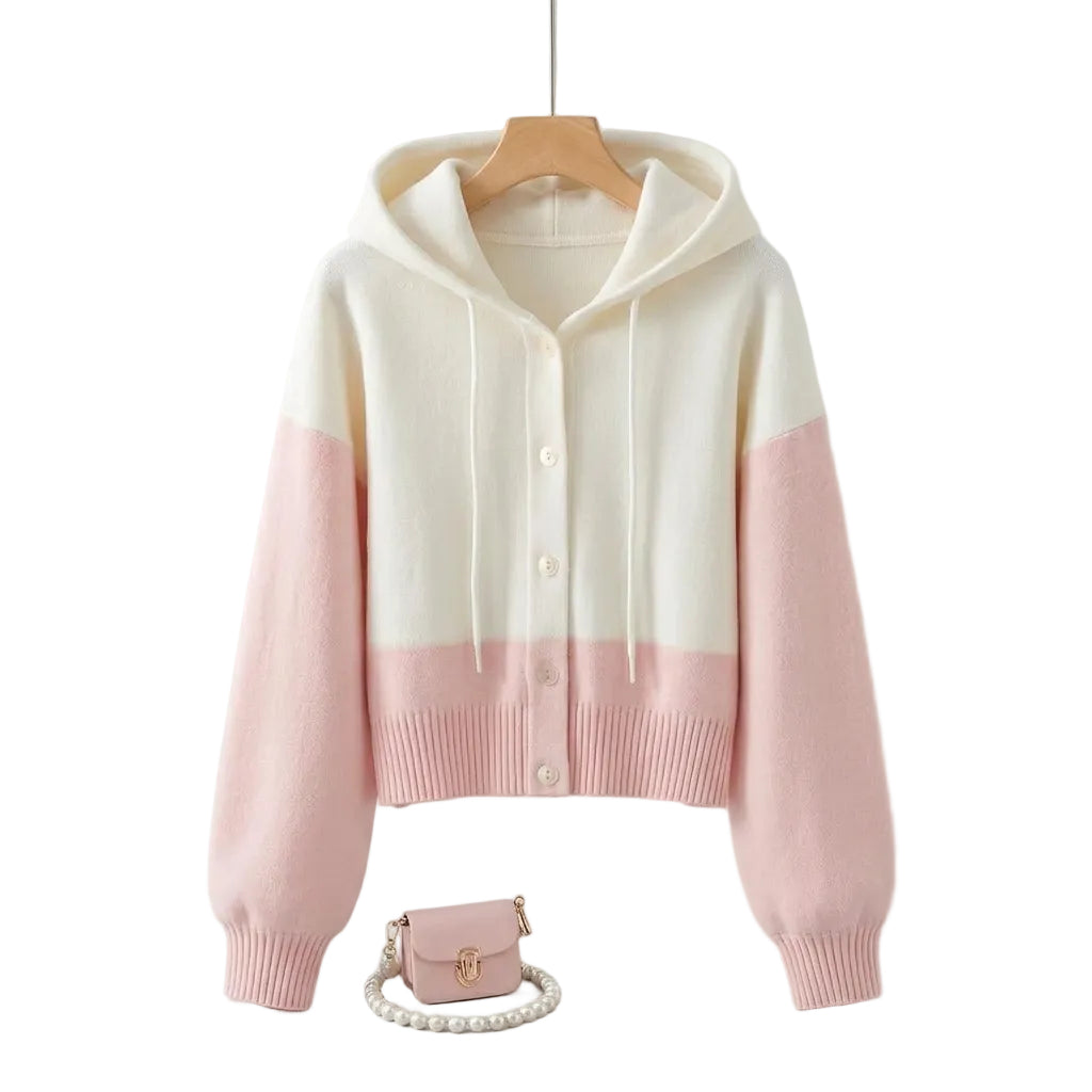 A color-blocked knit hoodie cardigan with a white upper body and hood and soft pink lower body and sleeves, styled on a hanger on a round table with a small pink purse and pearl necklace nearby.