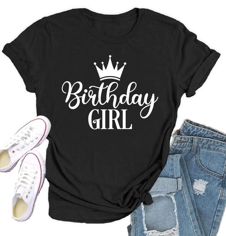 A black short-sleeve T-shirt with a white crown graphic and the words 'Birthday GIRL' printed on the front, styled in a flat lay with white sneakers and distressed blue jeans on a white background.
