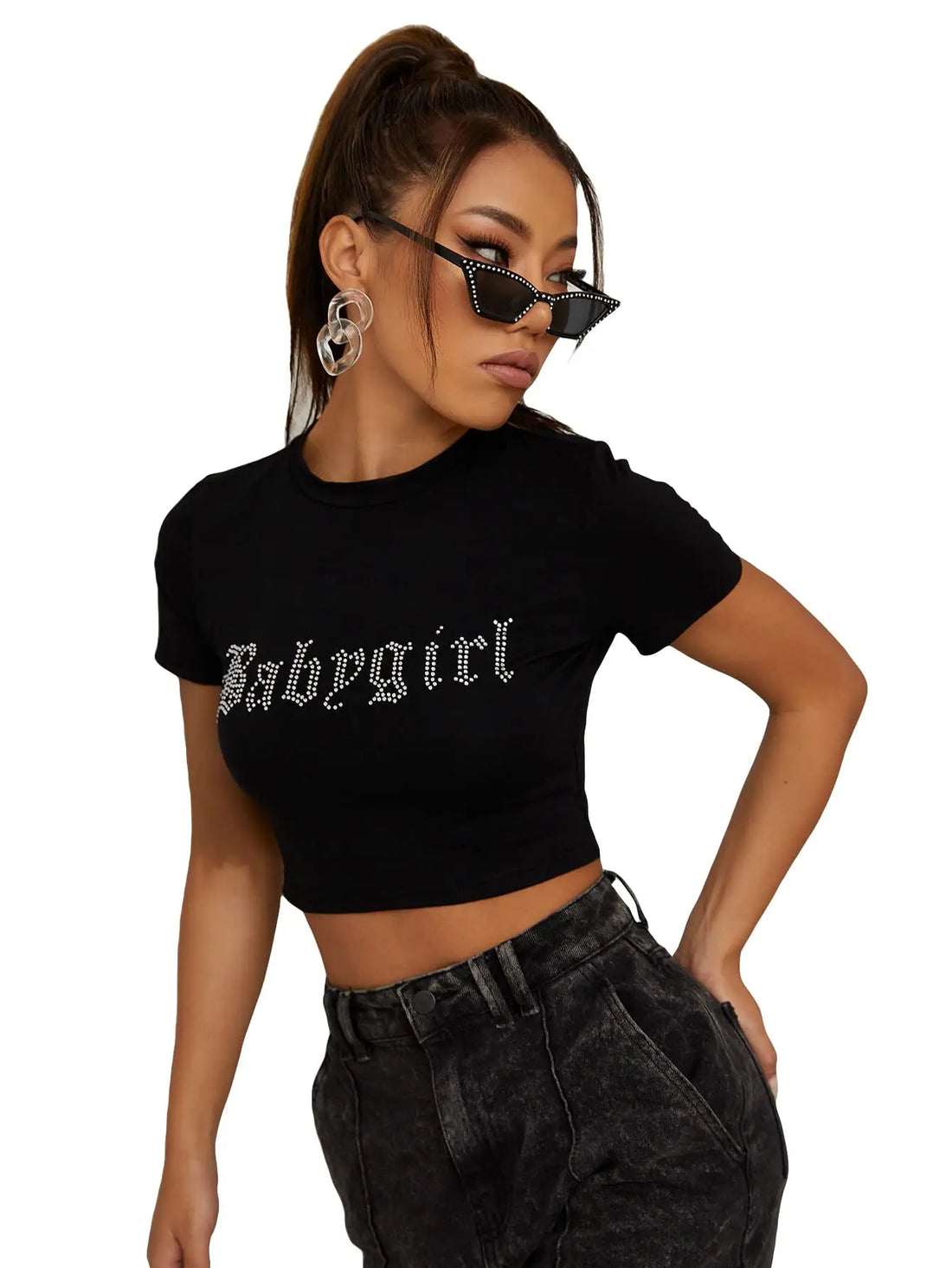 A woman modeling a black short-sleeve crop top with 'Babysirl' printed across the chest in a silver gothic font, paired with dark gray jeans and black sunglasses, against a plain white background.