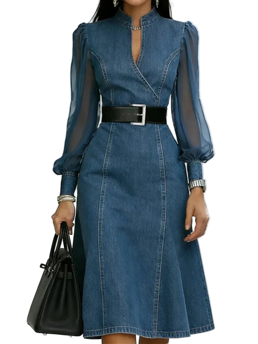 Blue denim dress with sheer sleeves and a black belt, held by a hand with a black bag on a white background.