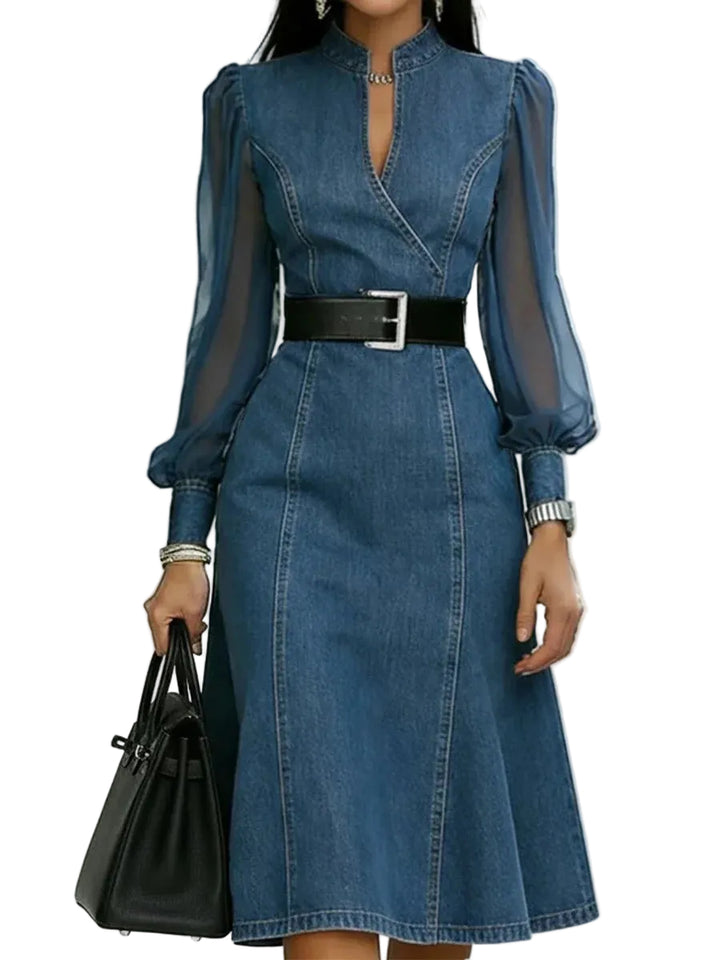 Blue denim dress with sheer sleeves and a black belt, held by a hand with a black bag on a white background.
