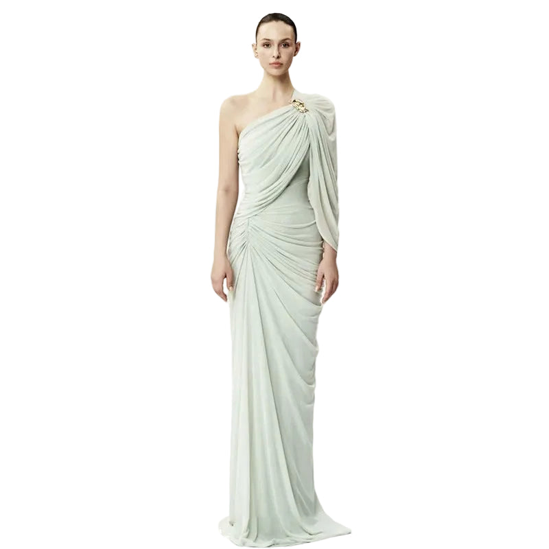 A model wears a floor-length, one-shoulder draped gown in a pale sage green with a small gold embellishment at the neckline, set against a plain white studio background.