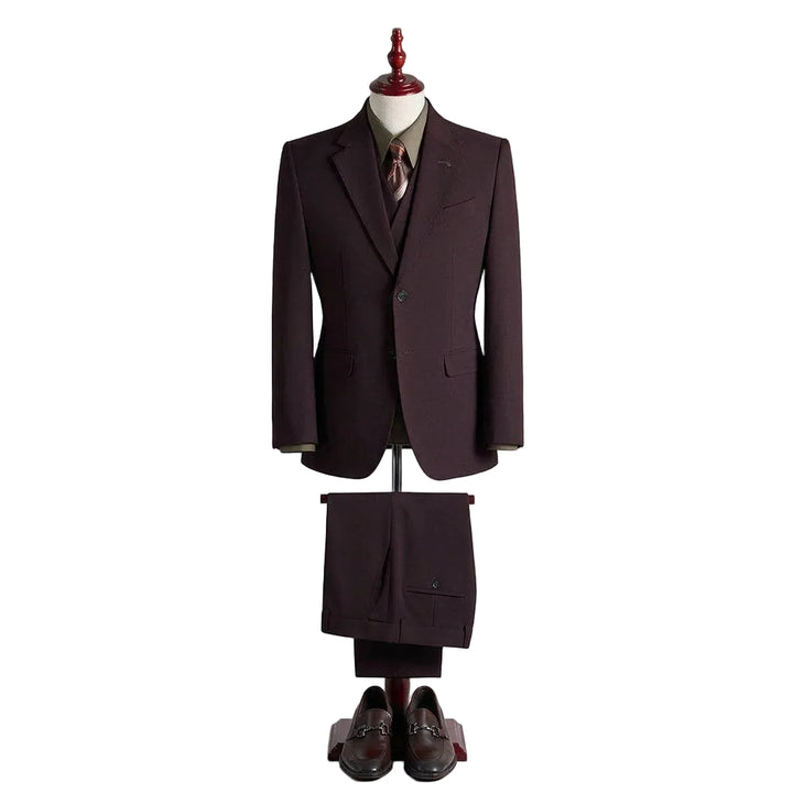 A dark plum-colored three-piece suit displayed on a mannequin, featuring a jacket, vest, trousers, a matching tie, and a pair of dark brown leather loafers, set against a neutral beige background.