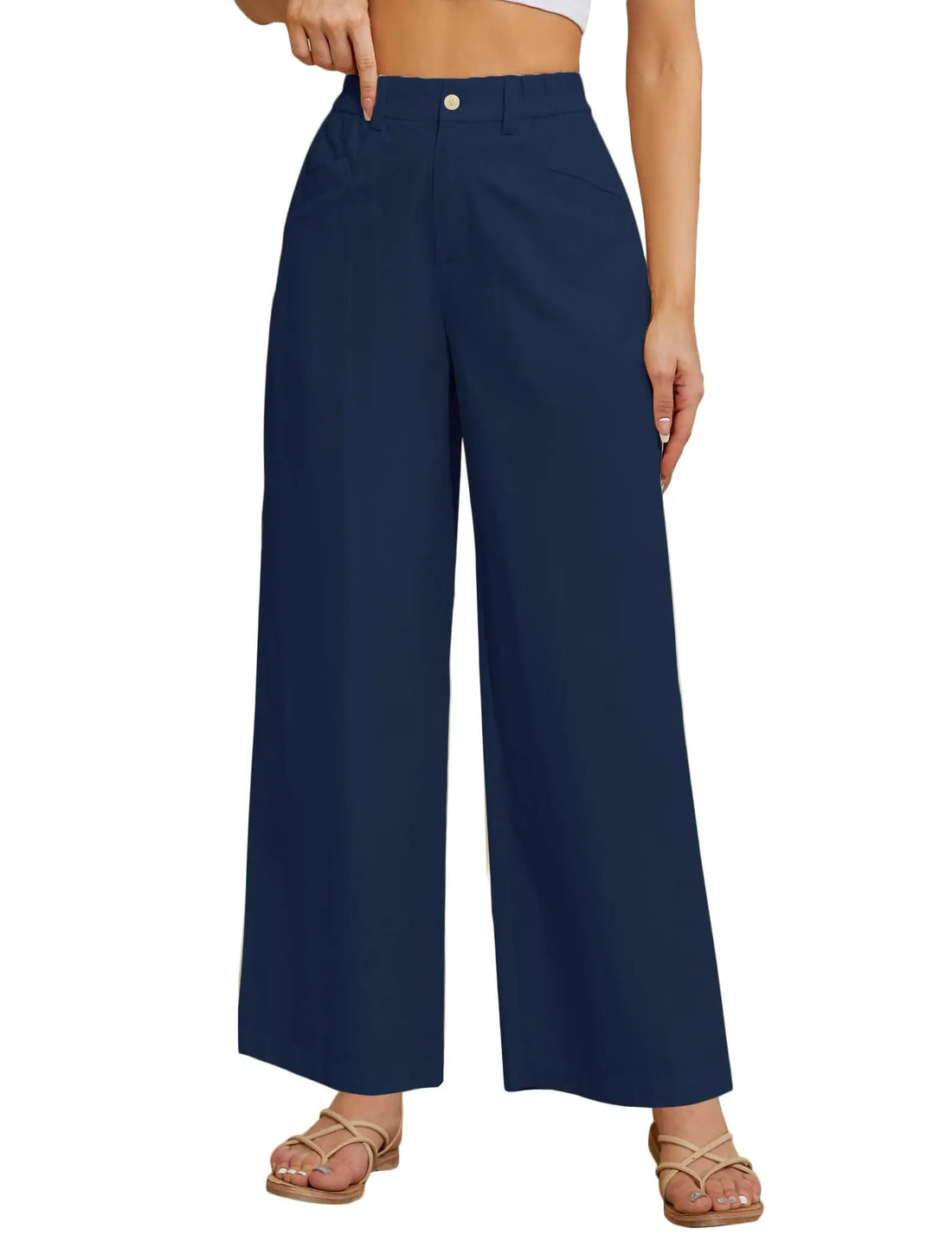 A model wearing a pair of high-waisted, wide-leg navy blue pants with a single gold button closure at the waistband, paired with beige strappy flat sandals against a plain white background.