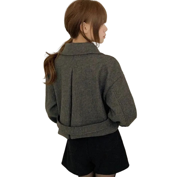 A woman shown from the back wearing a dark gray cropped coat with a wide collar and a rolled hem, paired with black shorts, against a plain off-white wall.