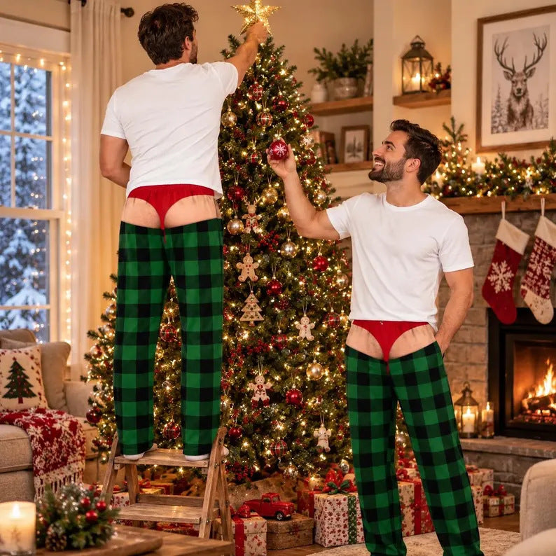 Two men in a cozy living room wearing green and black buffalo plaid pajama pants with red briefs, one standing on a ladder to hang a Christmas tree star while the other holds a ornament.