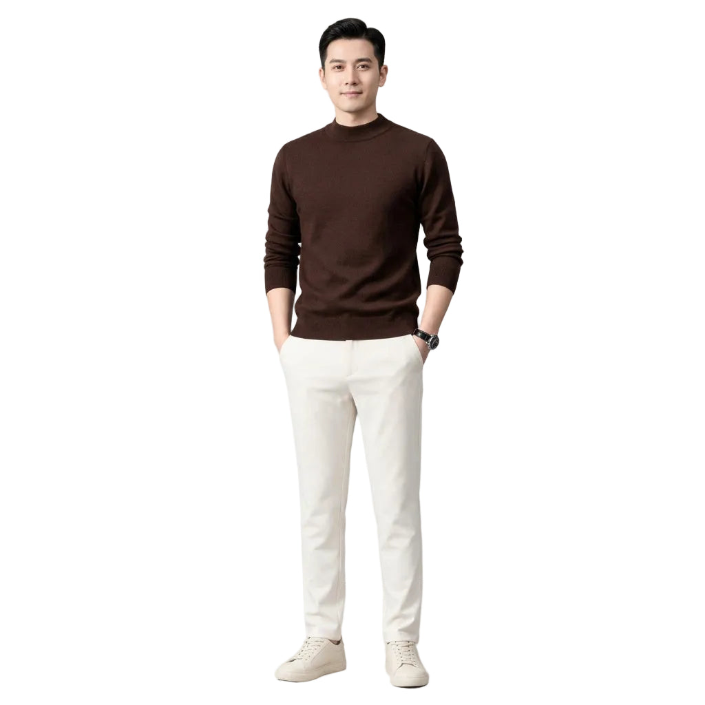 A man wearing a dark brown mock neck knit sweater and light beige trousers, standing against a dark gray background.