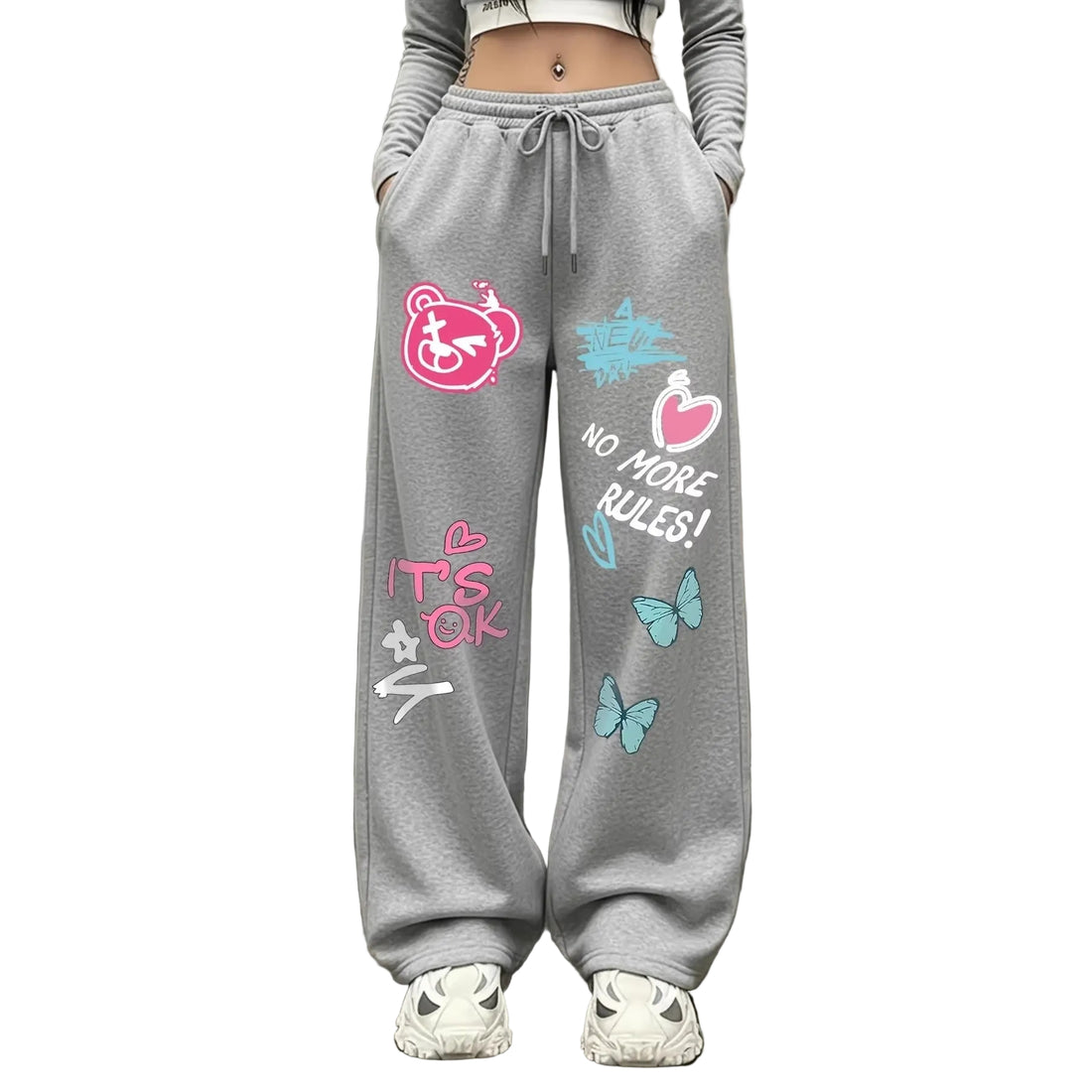 A person wearing gray drawstring sweatpants with colorful graphic prints, including a pink bear face, butterflies, hearts, and the text 'IT'S OK' and 'NO MORE RULES!'. The photo is cropped from the waist down, showing the pants paired with white sneakers against an indoor wall and floor background.