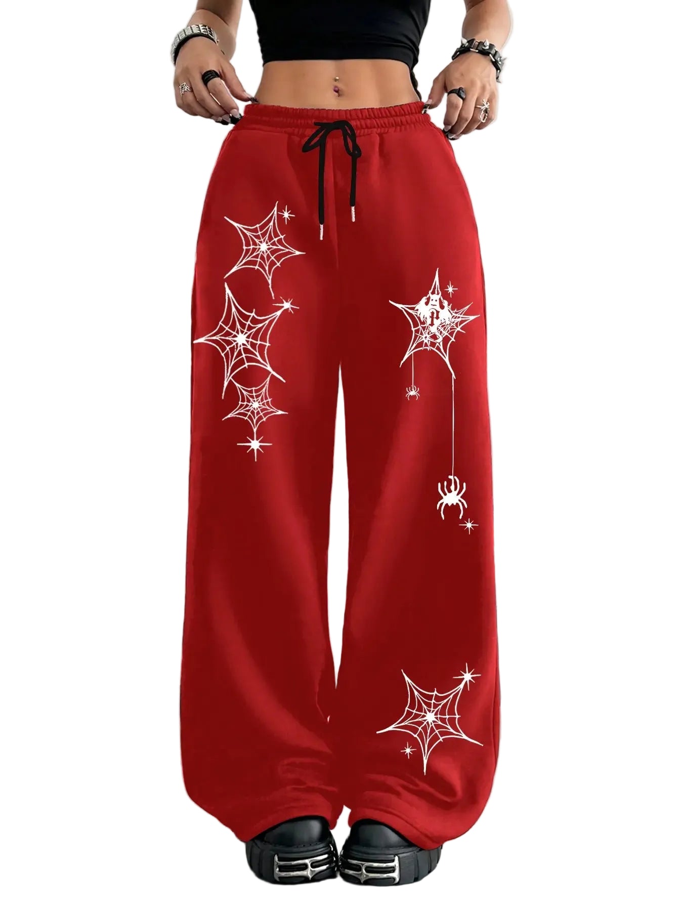 A person wearing bright red wide-leg sweatpants with white spiderweb and spider graphics, shown from the waist down in a styled indoor setting with Halloween decor. A vertical color swatch panel reading MORE COLORS appears on the left side.