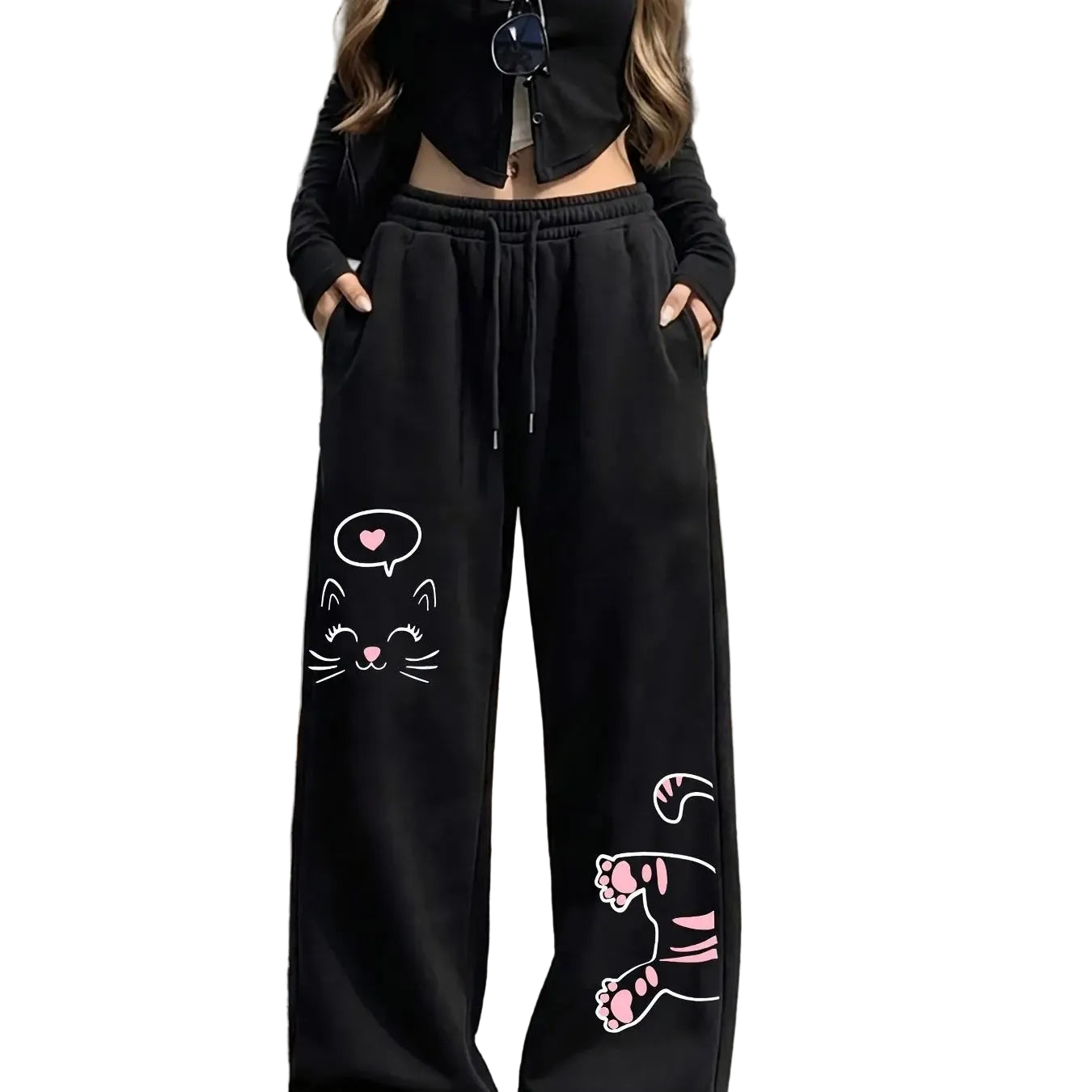 A person wearing black wide-leg sweatpants with pink and white cat face and paw prints, paired with a black cropped long-sleeve top, standing outdoors on a paved street.