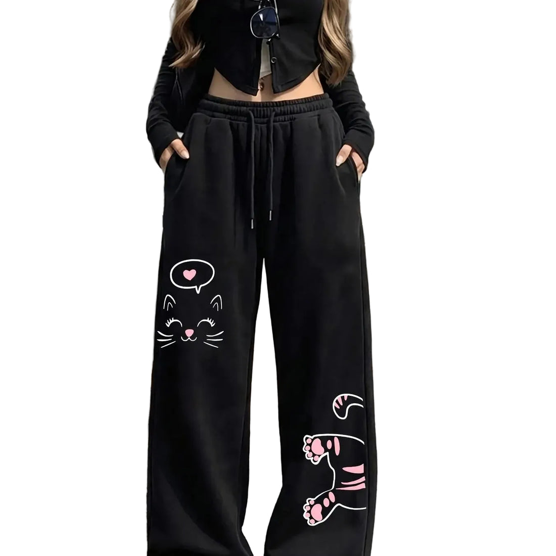 A person wearing black wide-leg sweatpants with pink and white cat face and paw prints, paired with a black cropped long-sleeve top, standing outdoors on a paved street.