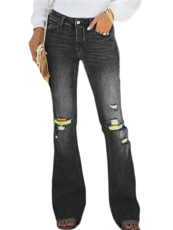 A person wearing dark gray distressed flare jeans with ripped details showing yellow lining, styled with a white long-sleeve top, brown sandals, and a small woven clutch on a plain white background.
